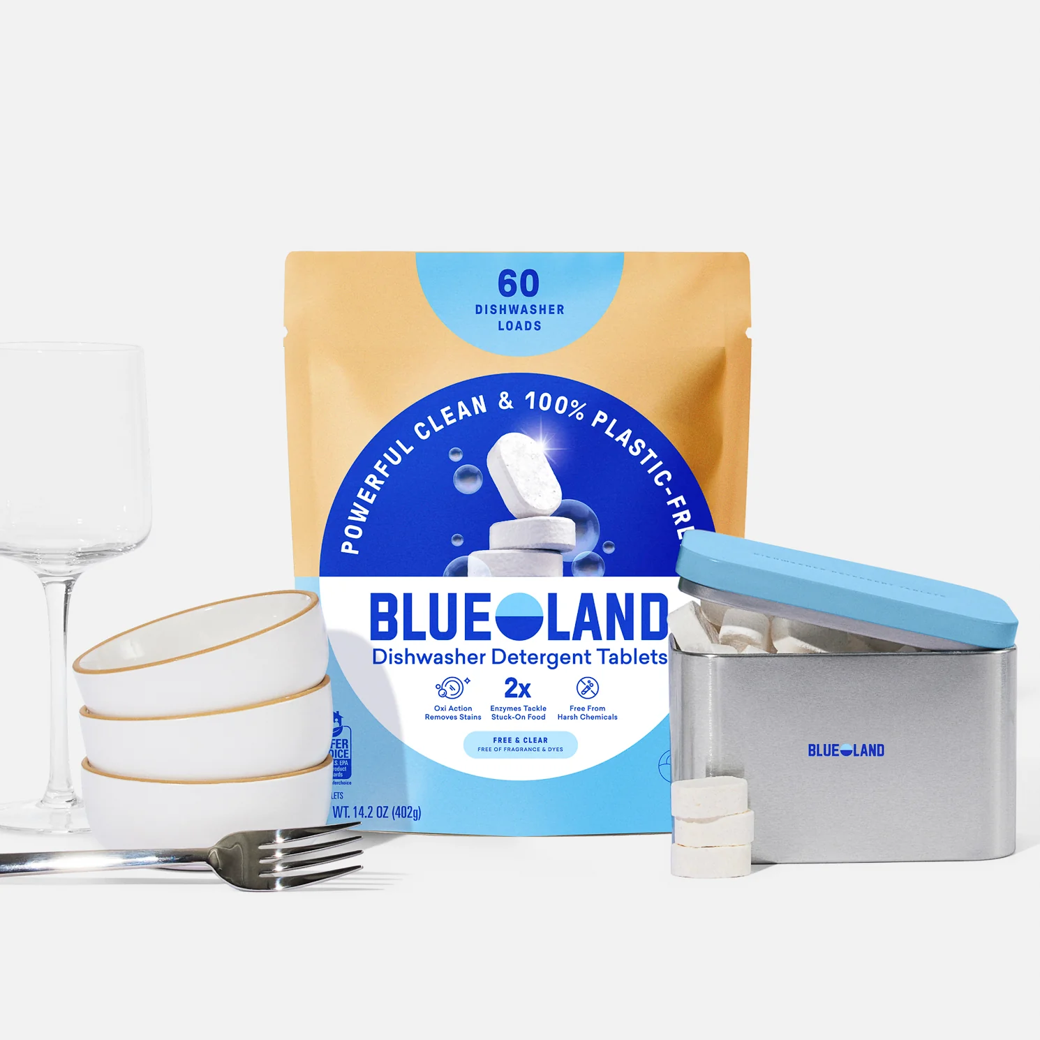A Dishwasher Detergent Starter Set sits behind a metal tin filled with tablets, beside a clear wine glass, stacked white dishes with gold rims, and silverware on a white surface.