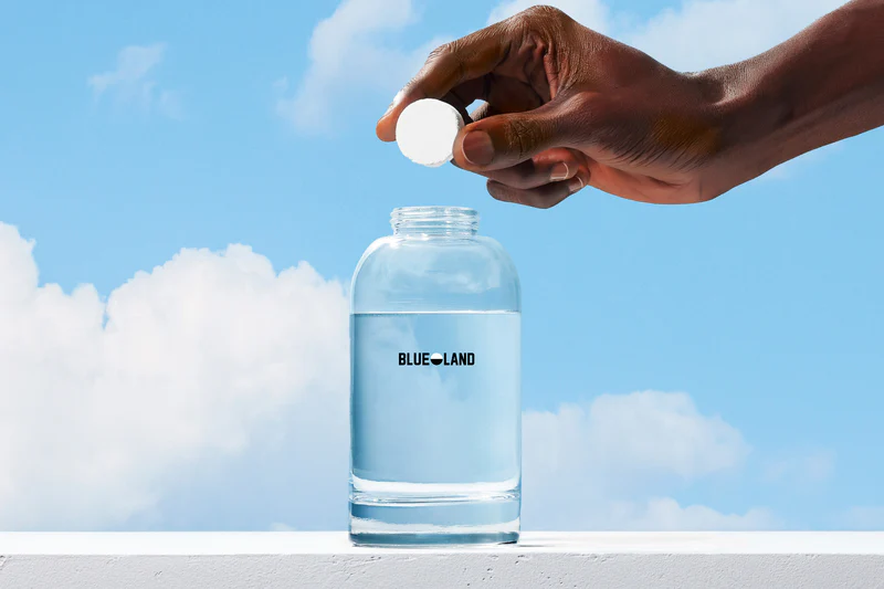 Hand holding a cleaning tablet above a Blue Land water bottle with a blue sky background